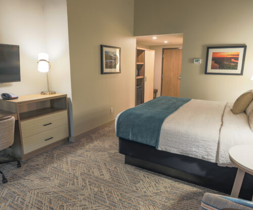 A guest room with one large bed. On one end of the room is a desk and TV. At the entrance of the room is a large wardrobe with a mini fridge. 