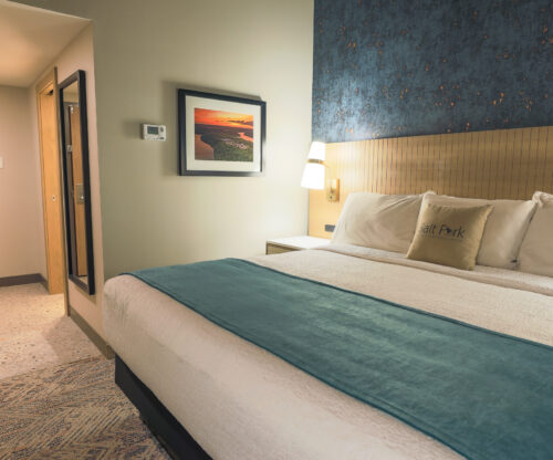 A guest room with a king bed. On the wall next to it is an image of Salt Fork Lodge at sunset. Leading to the bed area is the front entrance to the room and a body mirror on the wall.