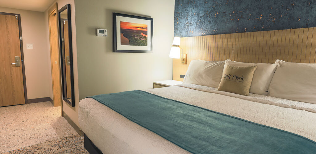 A guest room with a king bed. On the wall next to it is an image of Salt Fork Lodge at sunset. Leading to the bed area is the front entrance to the room and a body mirror on the wall.