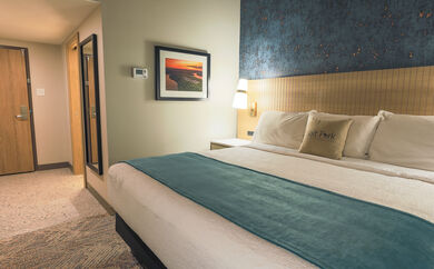 A guest room with a king bed. On the wall next to it is an image of Salt Fork Lodge at sunset. Leading to the bed area is the front entrance to the room and a body mirror on the wall.